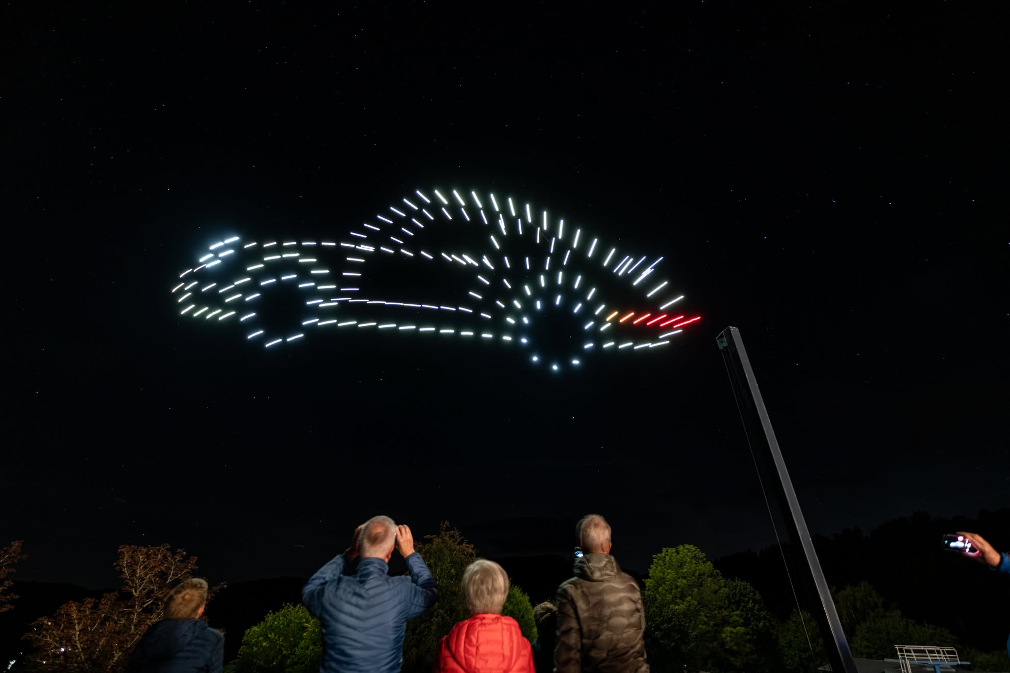 A Porsche 911 in white drones with a red tail light, over the crowd